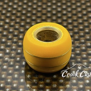 Puede incluir: Un objeto cilíndrico amarillo con un agujero central y dos líneas horizontales finas. El objeto tiene un acabado brillante y está sobre un fondo texturizado. El texto "Crook Customs" es visible en la esquina inferior derecha.