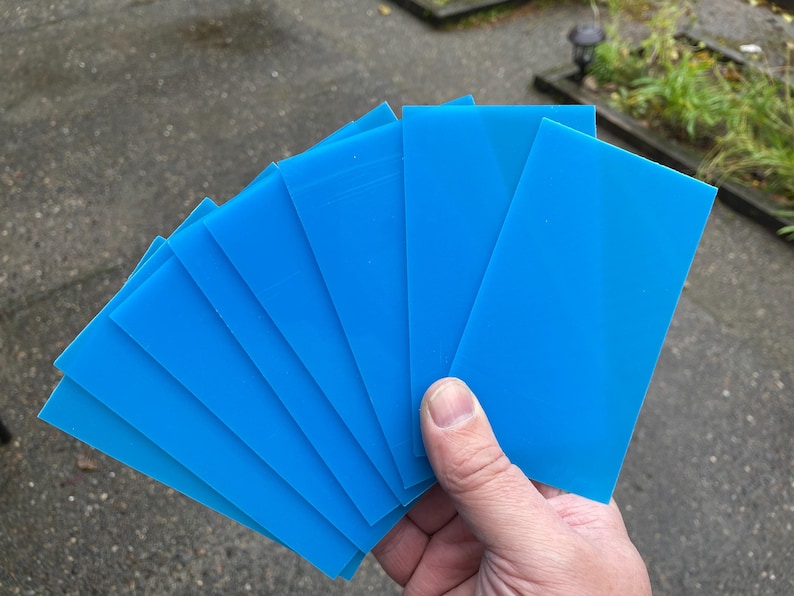 May include: A hand holds a fan of bright blue rectangular sheets. The sheets are evenly stacked, with a smooth, matte finish. The background is a blurred outdoor scene with concrete and greenery.