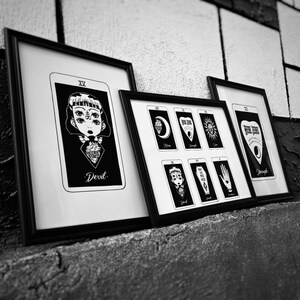 May include: Black and white framed tarot card art prints. The prints feature illustrations of tarot cards, including "Devil" and "Strength". The artwork has a gothic aesthetic, with detailed drawings and text.