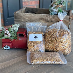 May include: Three clear plastic bags filled with pecan halves. The bags are sitting on a wooden surface. The largest bag is on the right, and the smallest bag is on the left. The middle bag is slightly larger than the left bag. The pecans are a light brown color and have a slightly shiny appearance.