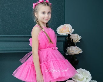 Puede incluir: Una niña pequeña con un vestido de tul rosa, una corona de flores y zapatillas blancas. Está posando sobre un cofre de terciopelo rosa con flores blancas en el fondo. El texto "Lily Land" es visible en el vestido.