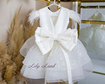 May include: White tulle dress with a large white satin bow, sequined bodice, and feather trim on the shoulders. The dress is hanging on a gold stand with a mirror in the background. The text "Lily Land" is visible on the dress.