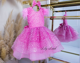 May include: A pink tulle dress with a sparkly bodice and feather sleeves. The dress has a layered skirt and a pink bow on the back. The dress is on a gold stand and the text "Lily Land" is visible in the bottom right corner.