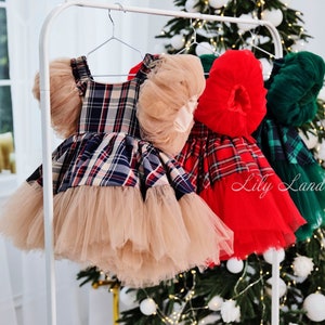 May include: Three plaid tulle dresses with ruffled sleeves hanging on a white rack. The dresses are red and black plaid, red and white plaid, and green and black plaid. The dresses are all on a white rack in front of a Christmas tree.