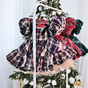 May include: Three plaid dresses with ruffled skirts and puffy sleeves hanging on a white rack in front of a Christmas tree with white ornaments.