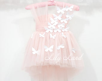 Puede incluir: Un vestido de tul rosa con aplicaciones de mariposas blancas. El vestido tiene un escote alto y un dobladillo alto-bajo. El vestido está en una percha y tiene el texto "Lily Land" en la parte inferior.
