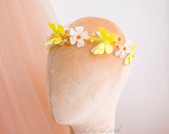 Può includere: Una corona di fiori gialla e bianca con farfalle e perline. La corona è su una testa di manichino.