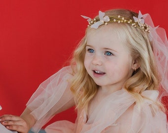 Può includere: Una bambina che indossa un abito di tulle pesca con pois bianchi e una corona di fiori. Sta tenendo una lanterna bianca con un ritaglio a forma di cuore. Lo sfondo è rosso pieno.