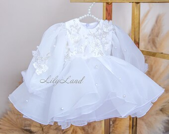 May include: A white tulle dress with pearl embellishments and long sleeves. The dress has a ruffled skirt and is hanging on a gold stand. The text "Lily Land" is visible on the dress.
