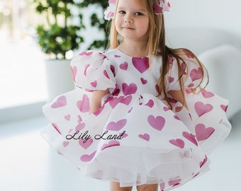 Puede incluir: Vestido blanco con estampado de corazones rosas y diadema a juego. El vestido tiene mangas abullonadas y una falda en capas. El texto "Lily Land" está impreso en el vestido. La niña lleva zapatos dorados.