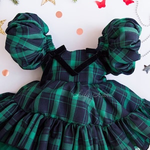 May include: A green and black plaid dress with puffed sleeves and a ruffled skirt. The dress has a black velvet bow at the neckline. The dress is on a white background with a few decorative items.