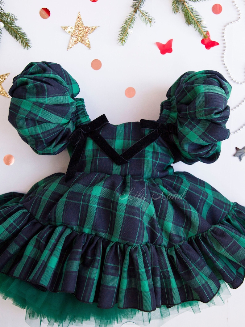 May include: A green and black plaid dress with puffed sleeves and a ruffled skirt. The dress has a black velvet bow at the neckline. The dress is on a white background with a few decorative items.