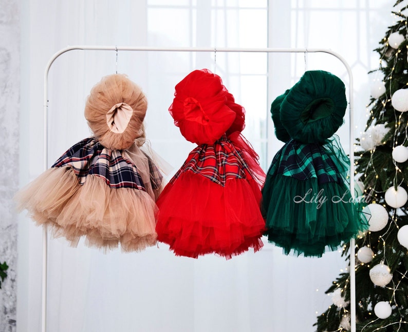 May include: Three tulle dresses with plaid accents hanging on a white rack. The dresses are beige, red, and green.