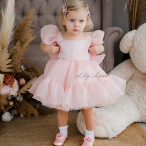 May include: A little girl wearing a pink tulle dress with puffed sleeves and a square neckline. She is standing on a rug with a white teddy bear in the background. The dress has a ruffled hem and the text "Lily Land" is visible on the dress.