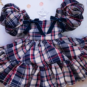 May include: A plaid dress with puffed sleeves and a ruffled skirt. The dress has a navy blue, red, and white plaid pattern and features blue velvet bows on the shoulders. The dress is layered over a tulle underskirt.