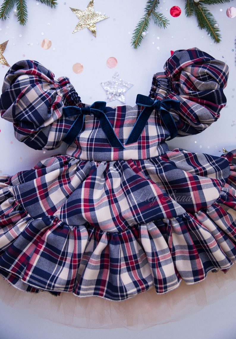 May include: A plaid dress with puffed sleeves and a ruffled skirt. The dress has a navy blue, red, and white plaid pattern and features blue velvet bows on the shoulders. The dress is layered over a tulle underskirt.