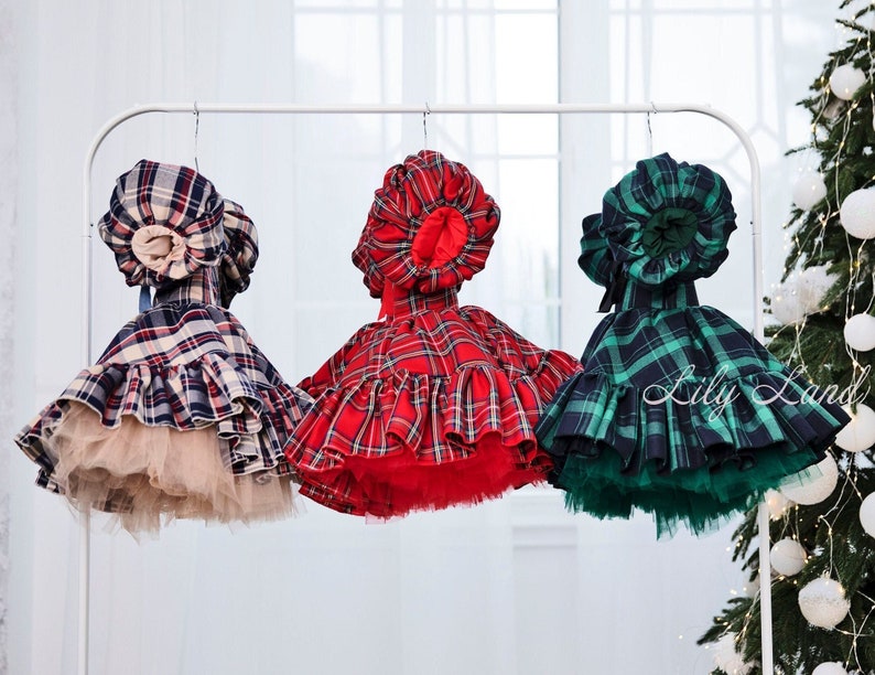 May include: Three plaid dresses with matching bonnets hanging on a white rack. The dresses are red, blue, and green with white plaid patterns. The bonnets are all white with a ruffled trim.