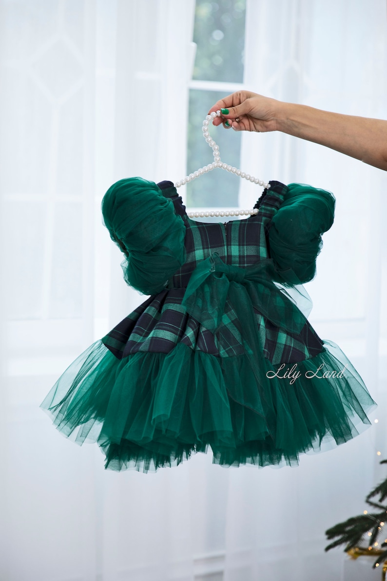 May include: A green plaid dress with a tulle skirt and puff sleeves. The dress is hanging on a white pearl hanger.