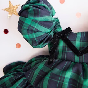 May include: A green and black plaid dress with a puffed sleeve and a black velvet bow. The dress has a ruffled skirt.