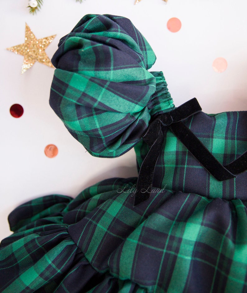 May include: A green and black plaid dress with a puffed sleeve and a black velvet bow. The dress has a ruffled skirt.