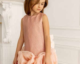 May include: A sleeveless, shimmering pink dress with a layered, rose-like skirt. The dress is worn by a young girl, and the overall style suggests a special occasion or formal wear. The background is a neutral white, emphasizing the dress's design.