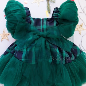 May include: Green plaid dress with a tulle skirt and puffed sleeves. The dress has a large green bow in the back. The dress is on a white background with green pine branches and gold stars.