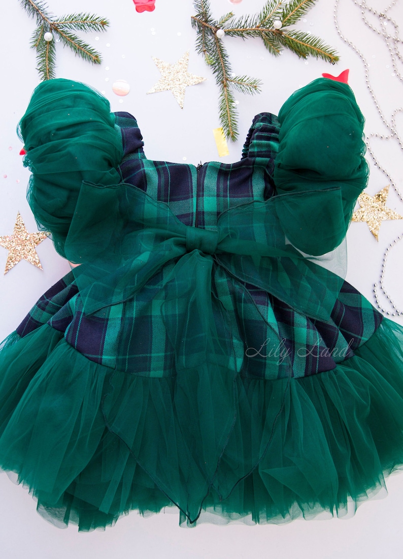 May include: Green plaid dress with a tulle skirt and puffed sleeves. The dress has a large green bow in the back. The dress is on a white background with green pine branches and gold stars.