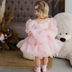 May include: A little girl wearing a pink tulle dress with a large bow in the back. The dress has puffed sleeves and a ruffled hem. The girl is standing in front of a brown armchair and a white teddy bear.