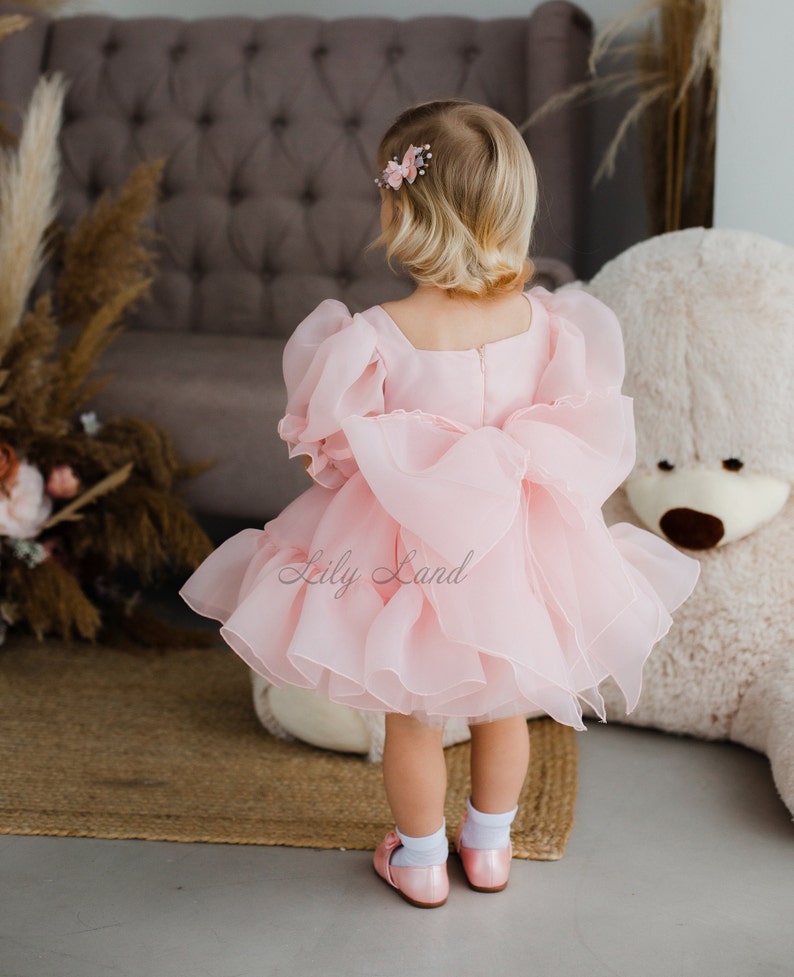 May include: A little girl wearing a pink tulle dress with a large bow in the back. The dress has puffed sleeves and a ruffled hem. The girl is standing in front of a brown armchair and a white teddy bear.