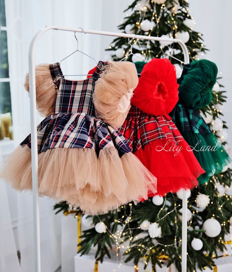 May include: Three plaid tulle dresses with ruffled sleeves hanging on a white rack. The dresses are red and black plaid, red and white plaid, and green and black plaid. The dresses are all on a white rack in front of a Christmas tree.