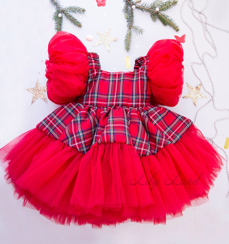 May include: Red plaid dress with a tulle skirt and puffy sleeves. The dress has a red and green plaid pattern and is decorated with a tulle skirt. The sleeves are puffy and made of a sheer fabric.