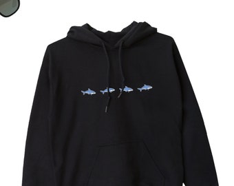 Puede incluir: Sudadera con capucha negra con una fila de cinco pequeñas ilustraciones de tiburones azules en la parte delantera.