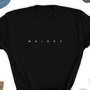 May include: A black t-shirt with the word "WHISKY" printed in white, centered across the chest. The shirt is a classic crew neck style, with short sleeves. The image also shows a pair of sunglasses, a hat, and shoes.