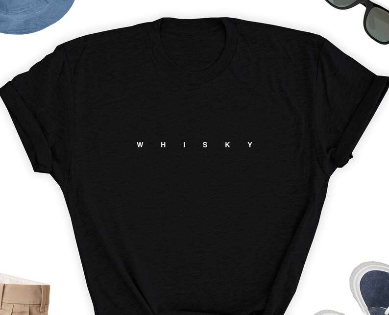 May include: A black t-shirt with the word "WHISKY" printed in white, centered across the chest. The shirt is a classic crew neck style, with short sleeves. The image also shows a pair of sunglasses, a hat, and shoes.