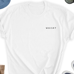 May include: A white t-shirt with the word "WHISKY" printed in black on the chest. The shirt has short sleeves and a crew neck. The image also shows a pair of sunglasses, a blue hat, and a pair of blue sneakers.