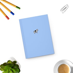 Puede incluir: Una libreta azul claro con una pegatina de vaca en blanco y negro en la portada.