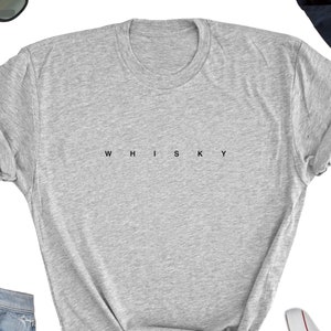 May include: A light gray t-shirt with the word "WHISKY" printed in black, centered across the chest. The shirt has a crew neck and short sleeves. Sunglasses, a dark blue hat, and a pair of jeans are visible in the background.