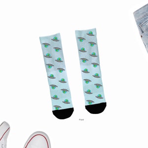 May include: A pair of light blue socks with a repeating pattern of cartoon aliens in flying saucers. The aliens have green heads and are in front of a blue planet. The socks have black heels and toes.