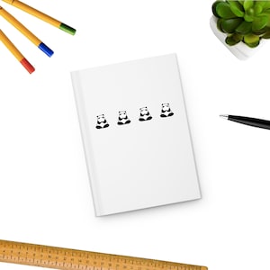 May include: A white notebook featuring four black and white panda illustrations. The notebook is on a white surface, with pens, a ruler, a pen, and a small plant in the background.