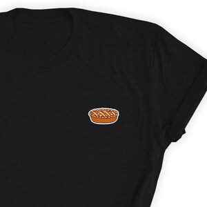 Puede incluir: Una camiseta negra con una pequeña imagen bordada de un pastel en el pecho izquierdo.
