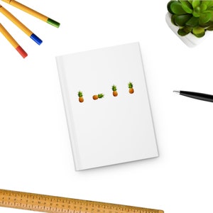 May include: A white notebook with a line of five pineapple emojis on a white surface. There are three yellow pencils, a blue pen, and a small potted succulent plant in the background.