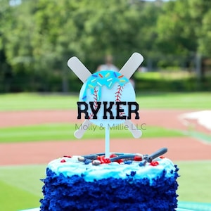 May include: A baseball-themed cake topper with the name "RYKER" in black block letters. The topper features a baseball with a blue donut design and crossed baseball bats. The cake is blue with a light blue frosting.