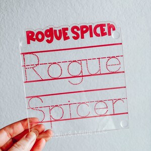Könnte beinhalten: Ein transparentes Kunststoff-Übungsboard mit roten gepunkteten Linien zum Üben des Schreibens des Namens "Rogue Spicer". Der Name "Rogue Spicer" ist oben auf dem Board in Rot gedruckt. Der Text "Molly & Millie LLC" ist unten auf dem Board in Rot gedruckt.