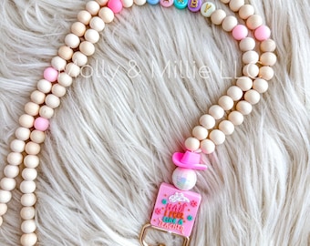 Puede incluir: Un cordón de cuentas rosa y blanco con un clip dorado y un dije rosa que dice "I may feel like a teacher". El cordón tiene el nombre "DARBY" escrito en cuentas de colores.