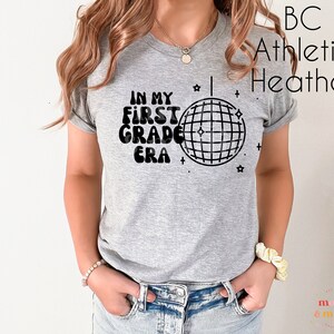 K&ouml;nnte beinhalten: Ein graues Heather-Athletik-T-Shirt mit schwarzem Text, der "In My First Grade Era" lautet, und einer Discokugel-Grafik.