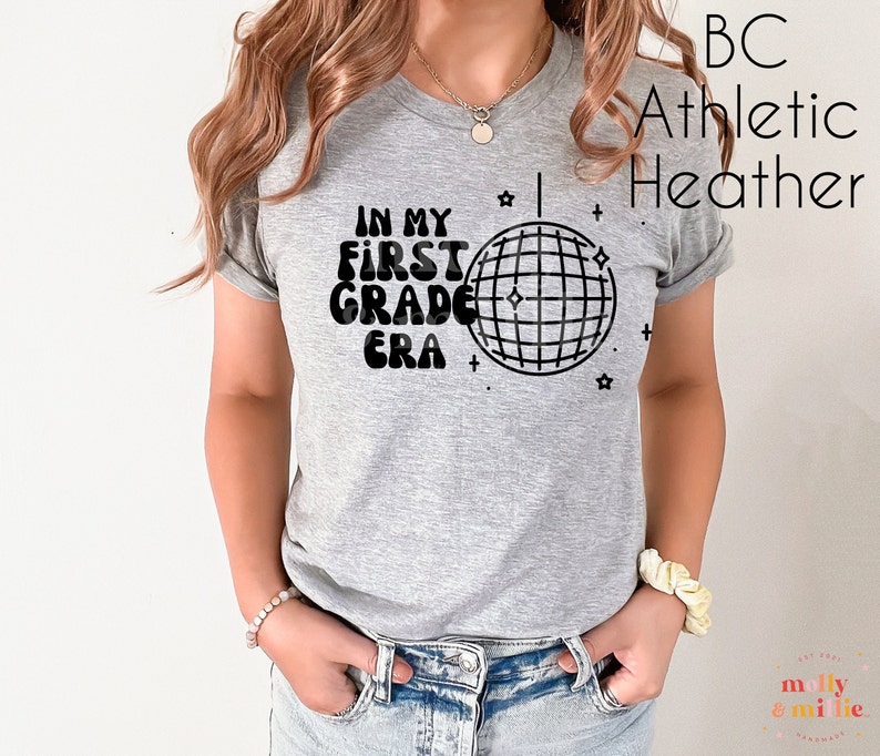 K&ouml;nnte beinhalten: Ein graues Heather-Athletik-T-Shirt mit schwarzem Text, der "In My First Grade Era" lautet, und einer Discokugel-Grafik.