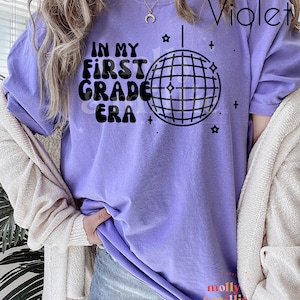 K&ouml;nnte beinhalten: Lilafarbenes T-Shirt mit dem Text "In My First Grade Era" und einer Discokugel-Grafik.