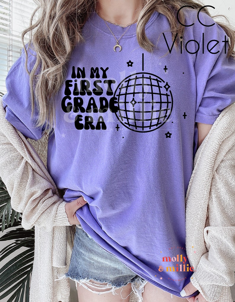K&ouml;nnte beinhalten: Lilafarbenes T-Shirt mit dem Text "In My First Grade Era" und einer Discokugel-Grafik.