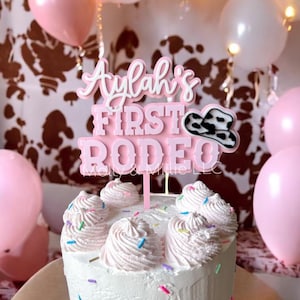 May include: A white frosted cake with pink frosting swirls and colorful sprinkles. A pink and white cake topper reads "Aylah's First Rodeo" with a cowboy hat graphic. Pink, white, and gold balloons are in the background.