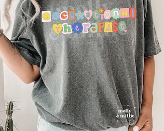 May include: A dark grey t-shirt with the text "Occupational Therapist" in colorful, outlined letters. The letters are decorated with smiley faces, hearts, and flowers.